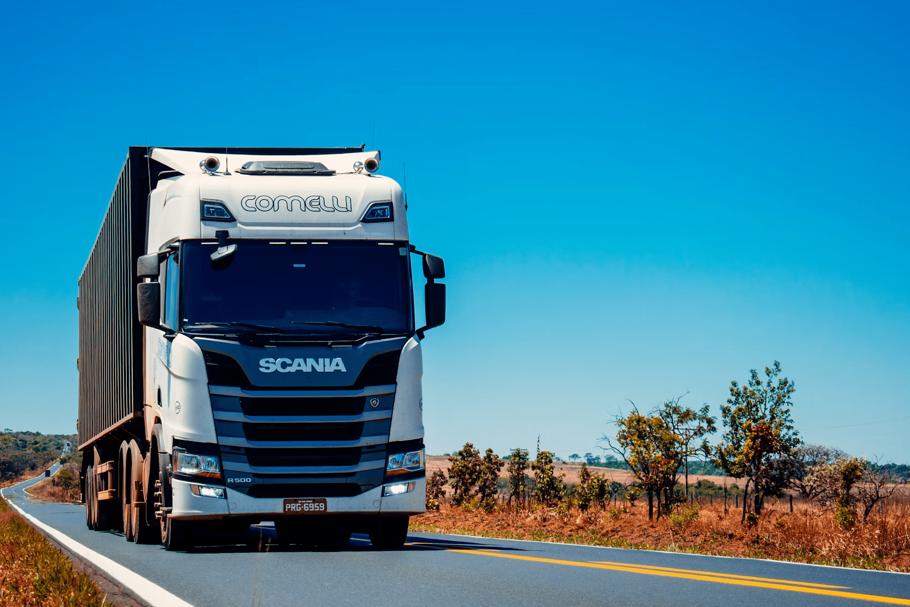 White Scania truck driving on a rural highway with clear blue skies in the background.